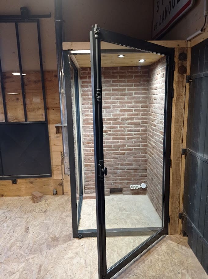 Fabrication et pose d'une porte et d'un châssis fixe vitrés pour une cave à vin personnalisée. Elle a une surface d'un mètre carré et une hauteur 2 mètres. Le mur du fond et celui côté droit sont habillés en briques jointées. Le côté gauche et constitué d'un châssis fixe en double vitrage. la porte est également en double vitrage dans un encadrement en tube à ailettes. Elle se verrouille à l'aide d'une crémone ferronée. L'ensemble et thermolaqué en ral 7016 gris. Cave à vin réalisée et installée par l'entreprise Claude Jeandin métallerie.