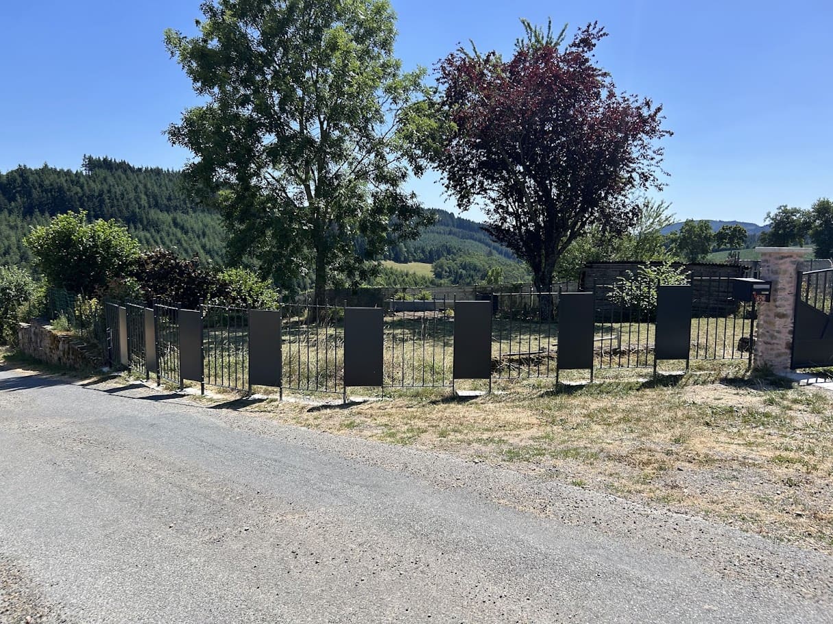 Fabrication et pose d'une clôture barreaudée pour une ancienne maison de campagne. Elle est constituée alternativement de section d'un mètre barreaudées verticalement et de sections de 50 cm tôlées. Les parties tôlées rappellent le design du portail et permettent de baisser le coût de l'ensemble de la clôture qui est longue de 47 ml. Deux sections encadrent le portail qui est fixés sur deux piliers en pierres maçonnés. L'ensemble est peint en ral 7016 gris. Clôture barreaudée métallique réalisée et installée par l'entreprise Claude Jeandin métallerie.