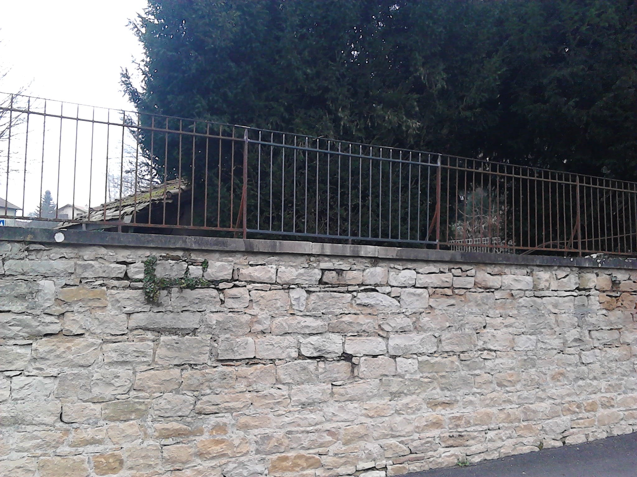 Restauration d'une clôture barreaudée en fer forgé sur un mur d'enceinte en pierres d'une propriété ancienne. Le barreaudage est verticale. L'extrémité de chaque barreaux rond et des poteaux carrés sont façonnés à la main en pointe de lance. Après évacuation de la section endommagée, la nouvelle section à été fixée par soudure sur site et elle s'intègre parfaitement à l'ensemble de la clôture qui n'a jamais été peinte. Clôture barreaudée métallique réalisée et installée par l'entreprise Claude Jeandin métallerie.
