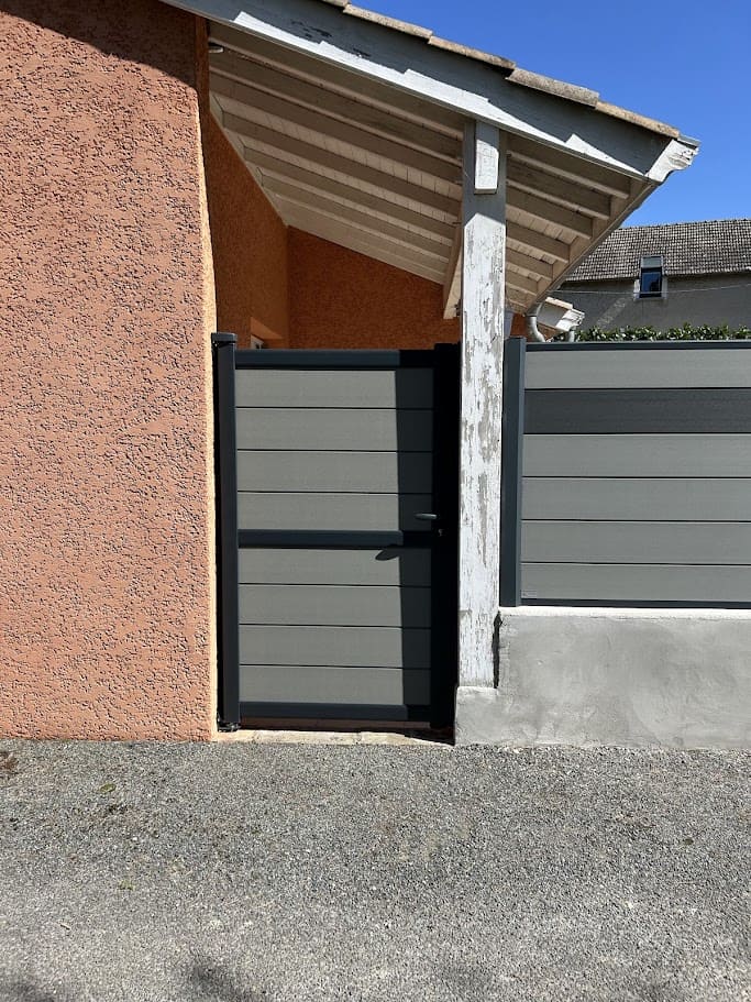 Pose d'un portillon et d'une clôture sur muret en composite de la marque Océwood. Les poteaux sont en aluminium ral 7016 et le remplissage composé de cinq rangées de lames décoratives horizontales teintes Basalte et d'une rangée intercalée de lames en teinte Graphite. La hauteur totale est de 180 cm. Portillon et clôture Océwood installés par l'entreprise Claude Jeandin métallerie.