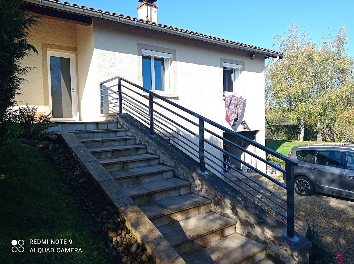 Fabrication et pose d'une rambarde métallique sur mesure pour un escalier extérieur. Le barreaudage est horizontale, il comprend deux sous lisses en tube rectangulaire et trois en plat, la main courante est en tube de section rectangulaire, les poteaux sont carrés. Elle est monobloc et mesure 6 mètres de long. L'ensemble est peint en gris ral 7016. Rambarde en fer réalisée par l'entreprise Claude Jeandin métallerie.