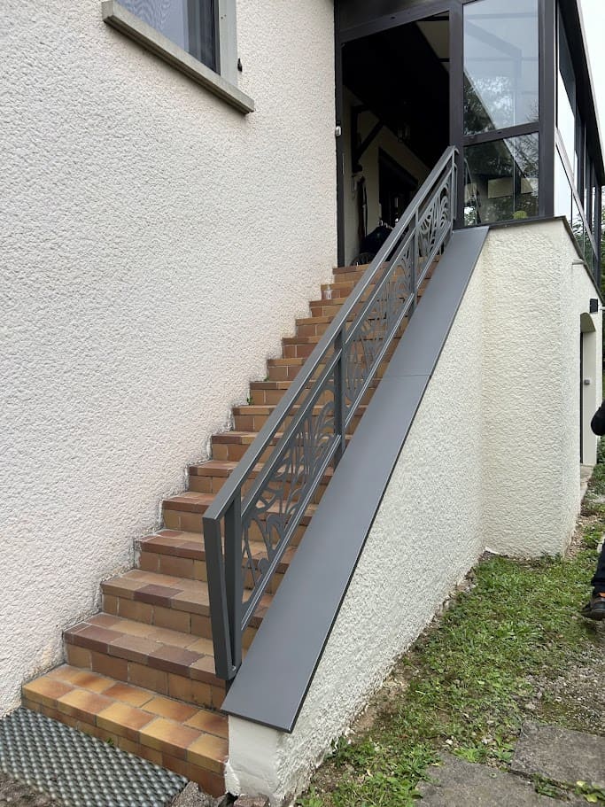 Fabrication et pose d'une rambarde métallique sur mesure pour un escalier extérieur. Elle est monobloc, constituée de trois panneaux en tôle à motifs découpés au laser. La main courante est en tube rectangulaire, les 4 poteaux de section carré. L'ensemble est peint en gris sablé ral 2900. Rambarde en fer design réalisée par l'entreprise Claude Jeandin métallerie.