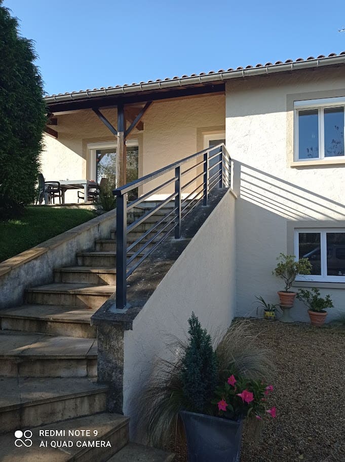 Fabrication et pose d'une rambarde métallique sur mesure pour un escalier extérieur. Le barreaudage est horizontale, il comprend deux sous lisses en tube rectangulaire et trois en plat, la main courante est en tube de section rectangulaire, les poteaux sont carrés. Elle est monobloc et mesure 6 mètres de long. L'ensemble est peint en gris ral 7016. Rambarde en fer réalisée par l'entreprise Claude Jeandin métallerie.