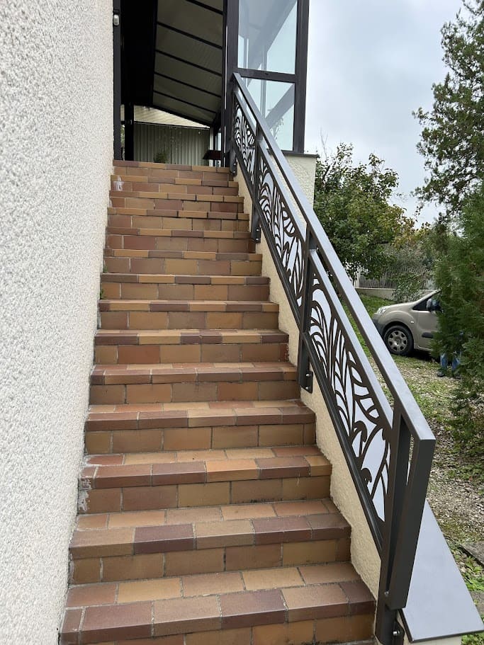 Fabrication et pose d'une rambarde métallique sur mesure pour un escalier extérieur. Elle est monobloc, constituée de trois panneaux en tôle à motifs découpés au laser. La main courante est en tube rectangulaire, les 4 poteaux de section carré. L'ensemble est peint en gris sablé ral 2900. Rambarde en fer design réalisée par l'entreprise Claude Jeandin métallerie.