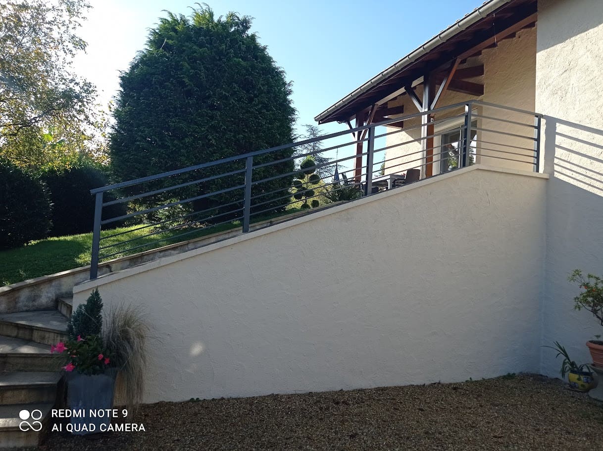 Fabrication et pose d'une rambarde métallique sur mesure pour un escalier extérieur. Le barreaudage est horizontale, il comprend deux sous lisses en tube rectangulaire et trois en plat, la main courante est en tube de section rectangulaire, les poteaux sont carrés. Elle est monobloc et mesure 6 mètres de long. L'ensemble est peint en gris ral 7016. Rambarde en fer réalisée par l'entreprise Claude Jeandin métallerie.